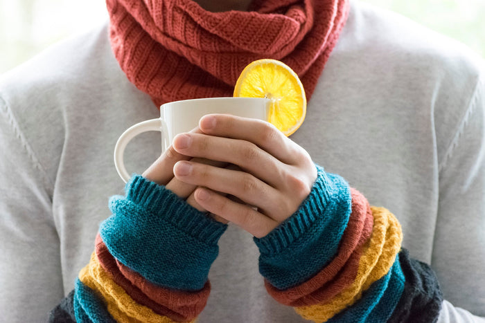 Eine junge Frau mit Schal und Armwärmern hält einen heißen Becher Tee mit Zitrone.
