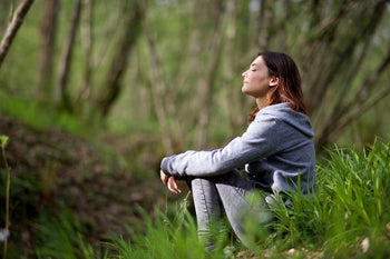 Eine junge Dame sitzt im Wald auf dem Rasen und genißet die Sonne. Sie hat Ihre Augen geschlossen und versucht zu entspannen.