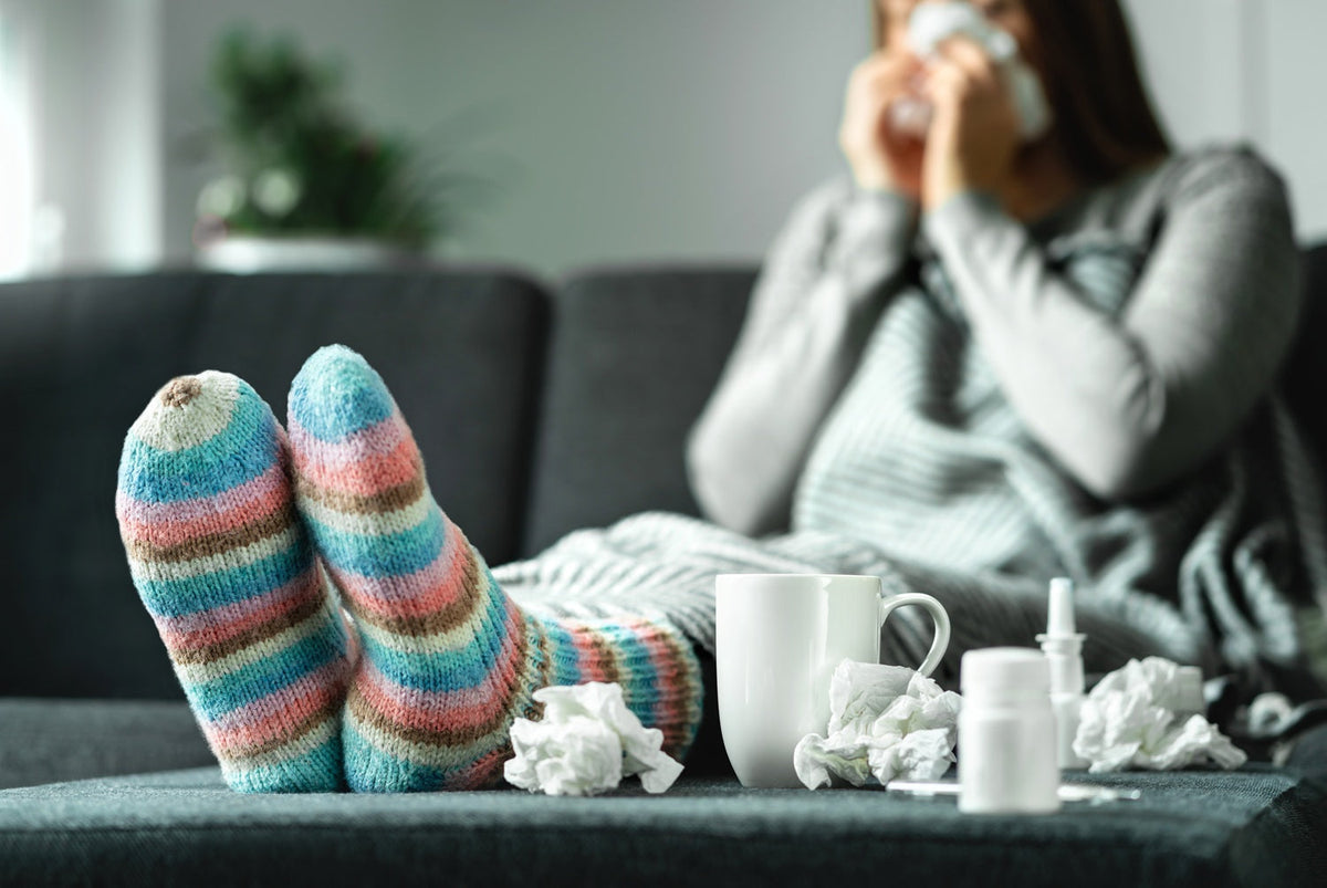 Eine junge Frau sitzt in dicken Klamotten mit einer Decke auf der Couch. Vor ihr liegen Nasenspray, gebrauchte Taschentücher und eine Tasse mit Tee. Sie ist erkältet.