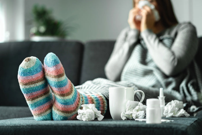 Eine junge Frau sitzt in dicken Klamotten mit einer Decke auf der Couch. Vor ihr liegen Nasenspray, gebrauchte Taschentücher und eine Tasse mit Tee. Sie ist erkältet.